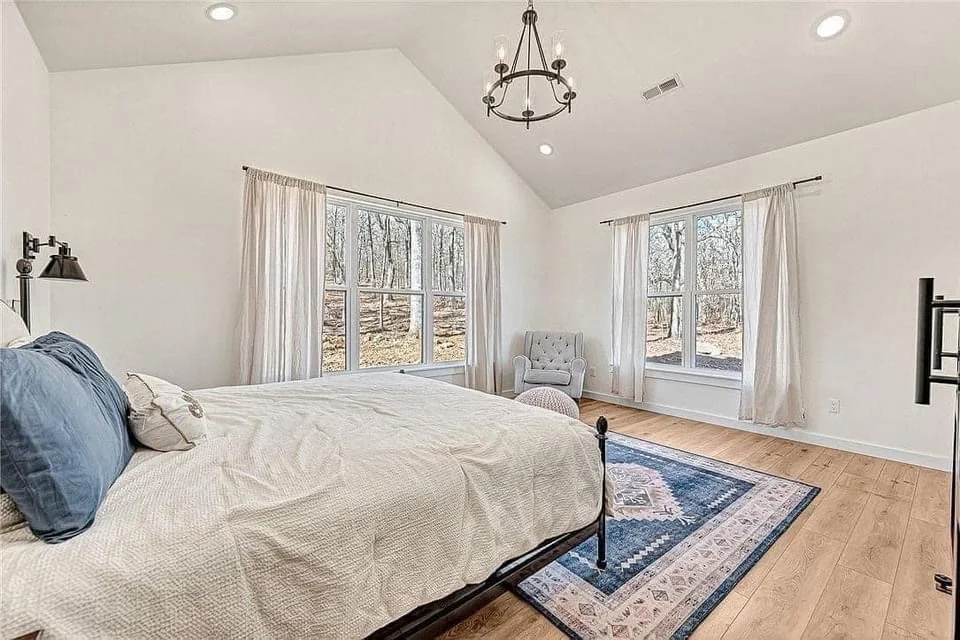 Durable Home Renovations In Troy, MO | BECC Builders, LLC A white bedroom with a bed, hardwood flooring, a blue rug, a black chandelier, and large windows with the curtains open.