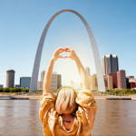 A woman with her hands in the air, making the heart symbol in front of the St. Louis Gateway Arch.
