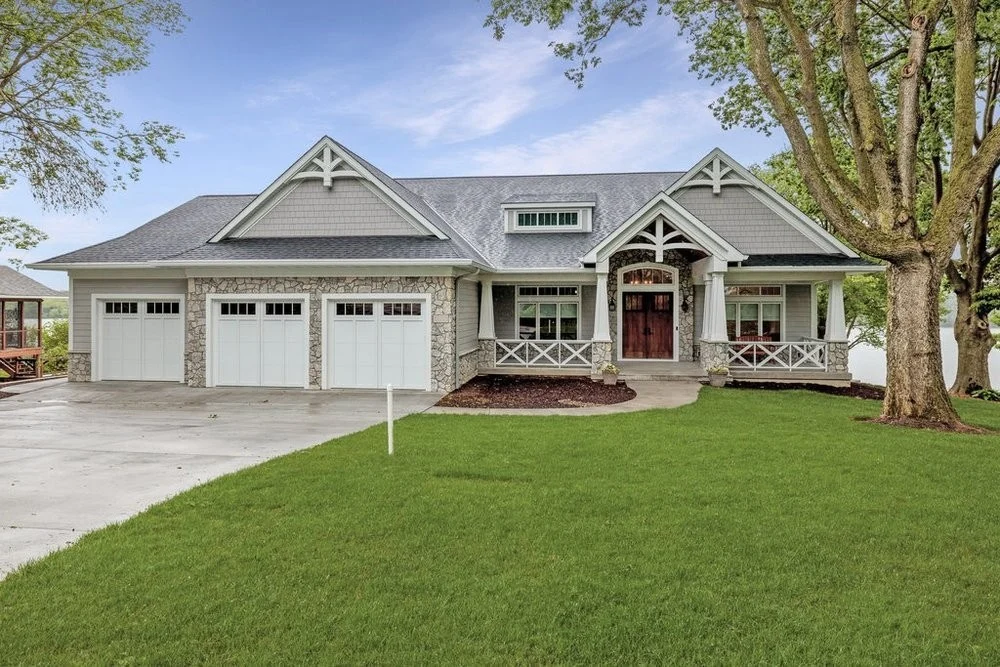 Elegant custom home with stone accents, three-car garage, covered porch, large front lawn, mature tree, and gray shingle roof.