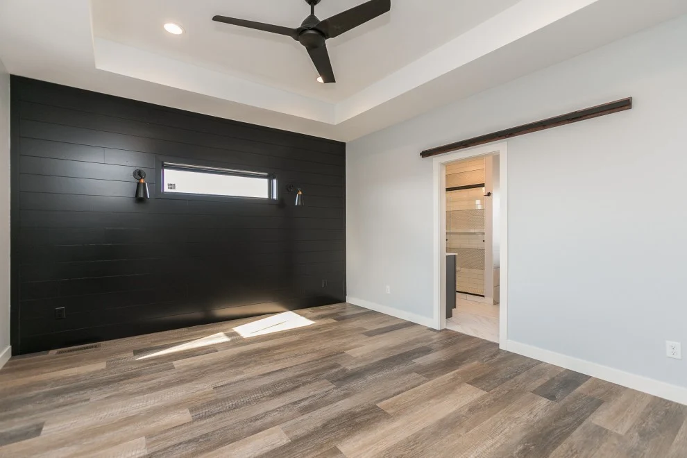 Finished home addition room with black accent wall, wood-look flooring, tray ceiling, ceiling fan, recessed lights, and bathroom entry.