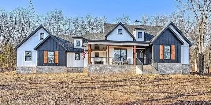 Modern custom farmhouse with white siding, dark gables, covered porch, stone foundation, and wooded lot under a clear blue sky.