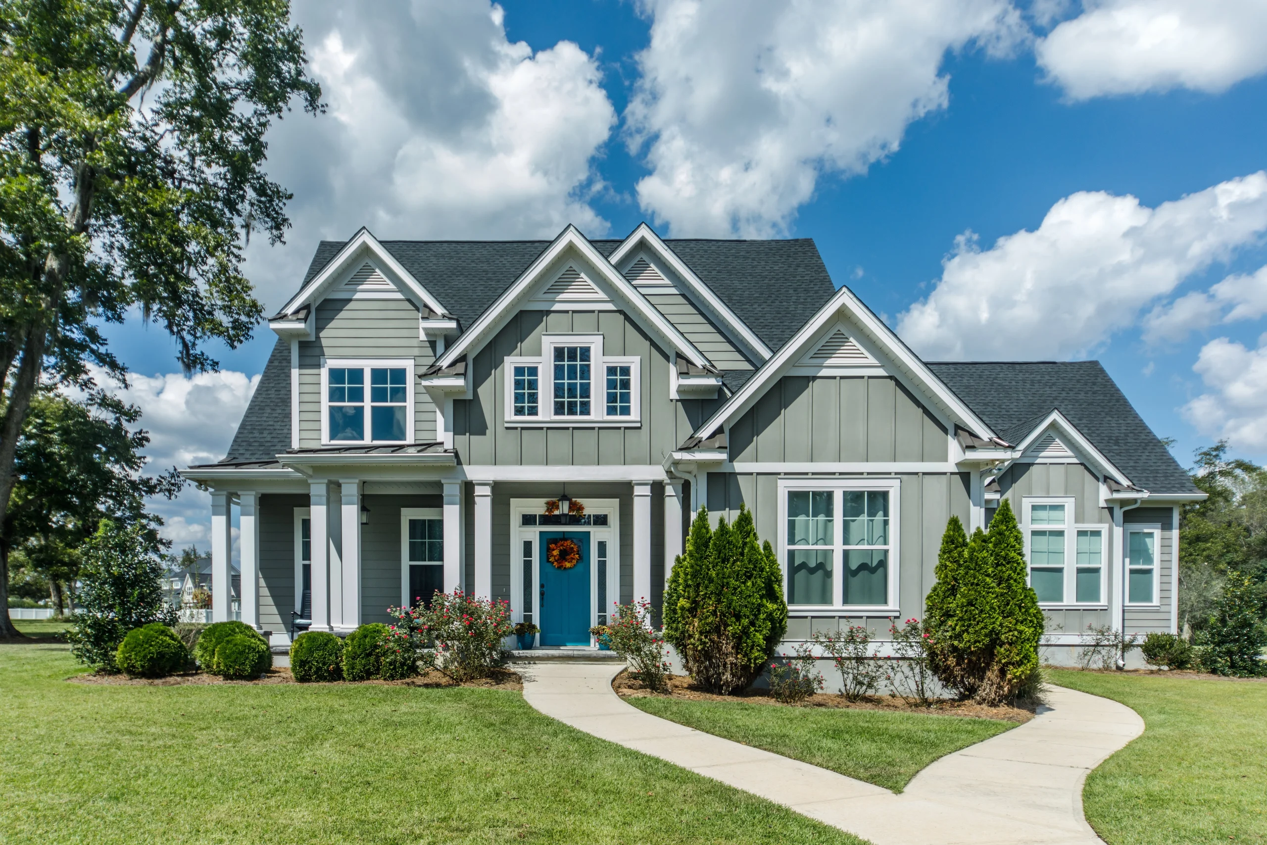 Gray custom home with blue front door, gabled rooflines, white trim, curved walkway, landscaped shrubs, and bright cloudy sky.