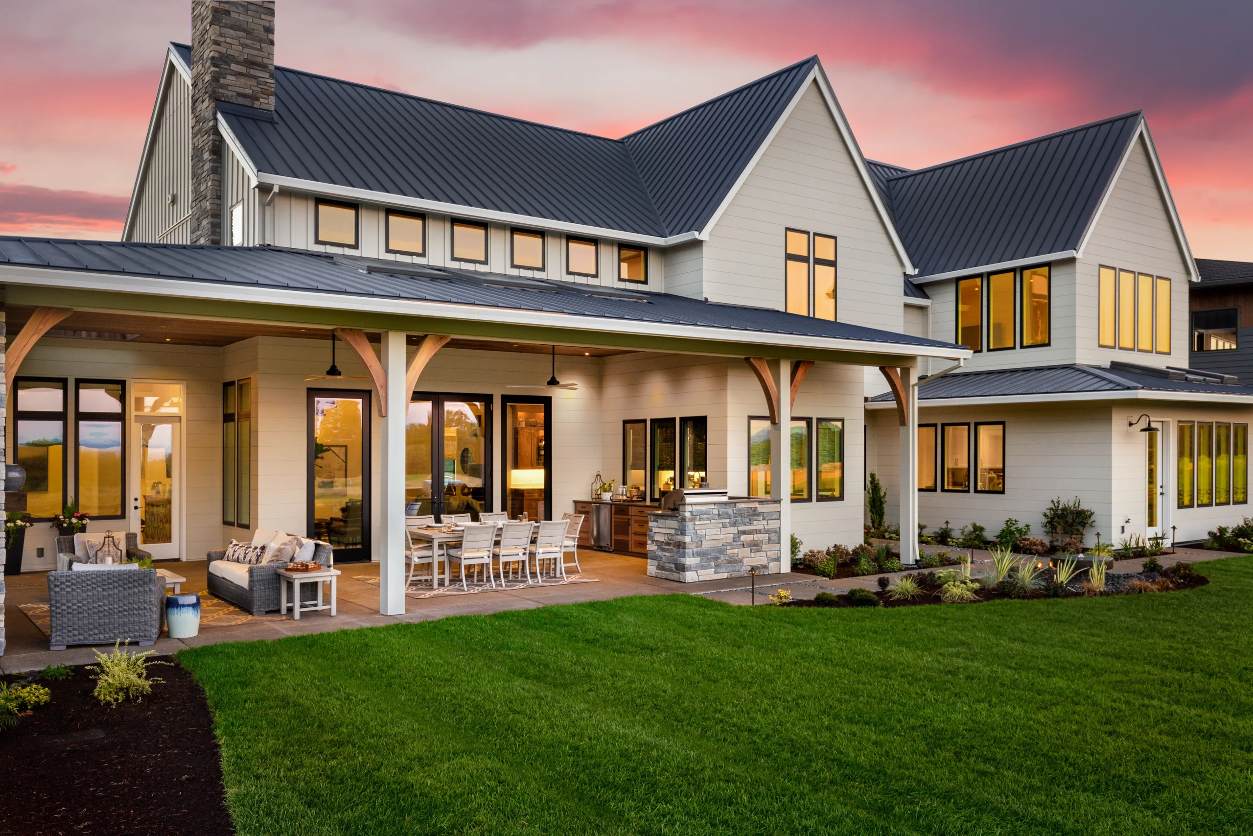 Custom home with covered outdoor living patio, dining area, black metal roof, warm windows, stone accents, and sunset sky.