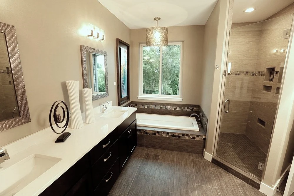 A modern bathroom with a glass-enclosed, step-in shower in the back right corner, a rectangular tub along the back wall, and a double-sink vanity with black cabinets and a white countertop on the left. There is a tall rectangular window over the tub.