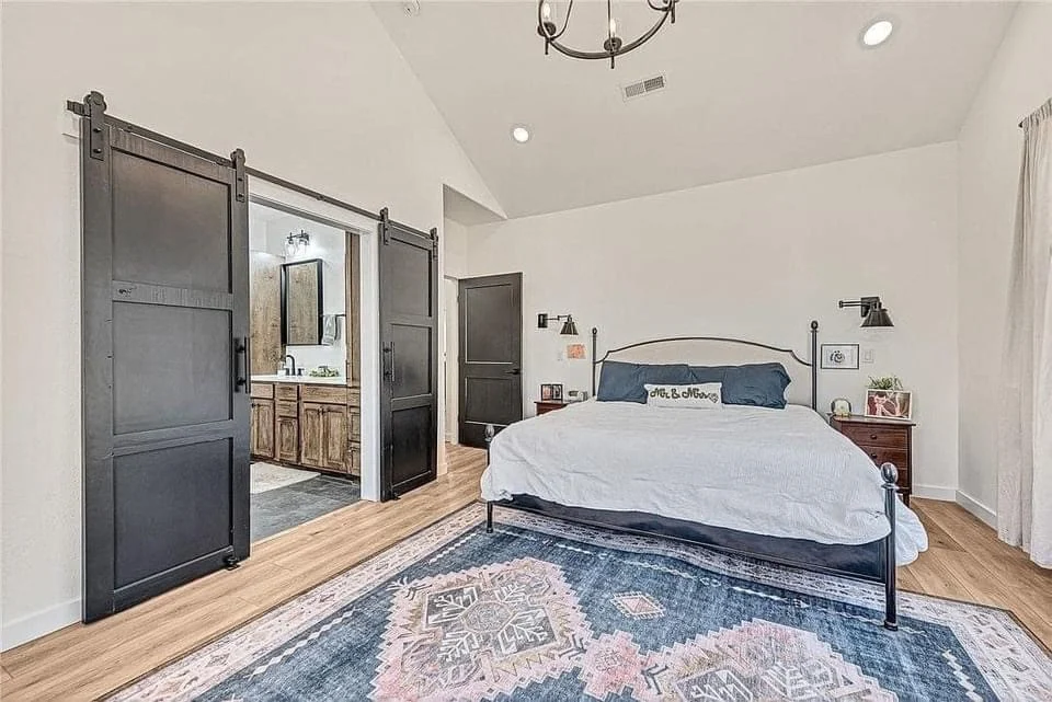 A bedroom with high ceilings and a low, wide bed. There is a set of black sliding farmhouse doors on the left, partially opened to reveal the bathroom. The flooring is light colored wood, the walls are cream, and there is a blue, cream, and white area rug on the floor beneath the bed.