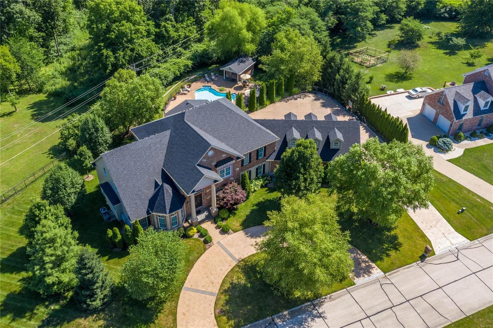 A photo taken from above of a large, multi-story red brick home with a gray shingled roof. Parts of a semi-circular driveway can be seen in the front of the house, and a larger parking area is visible in the back. There's a small, disconnected structure next to an in-ground pool in the backyard. The house is surrounded by trees and bushes for privacy.