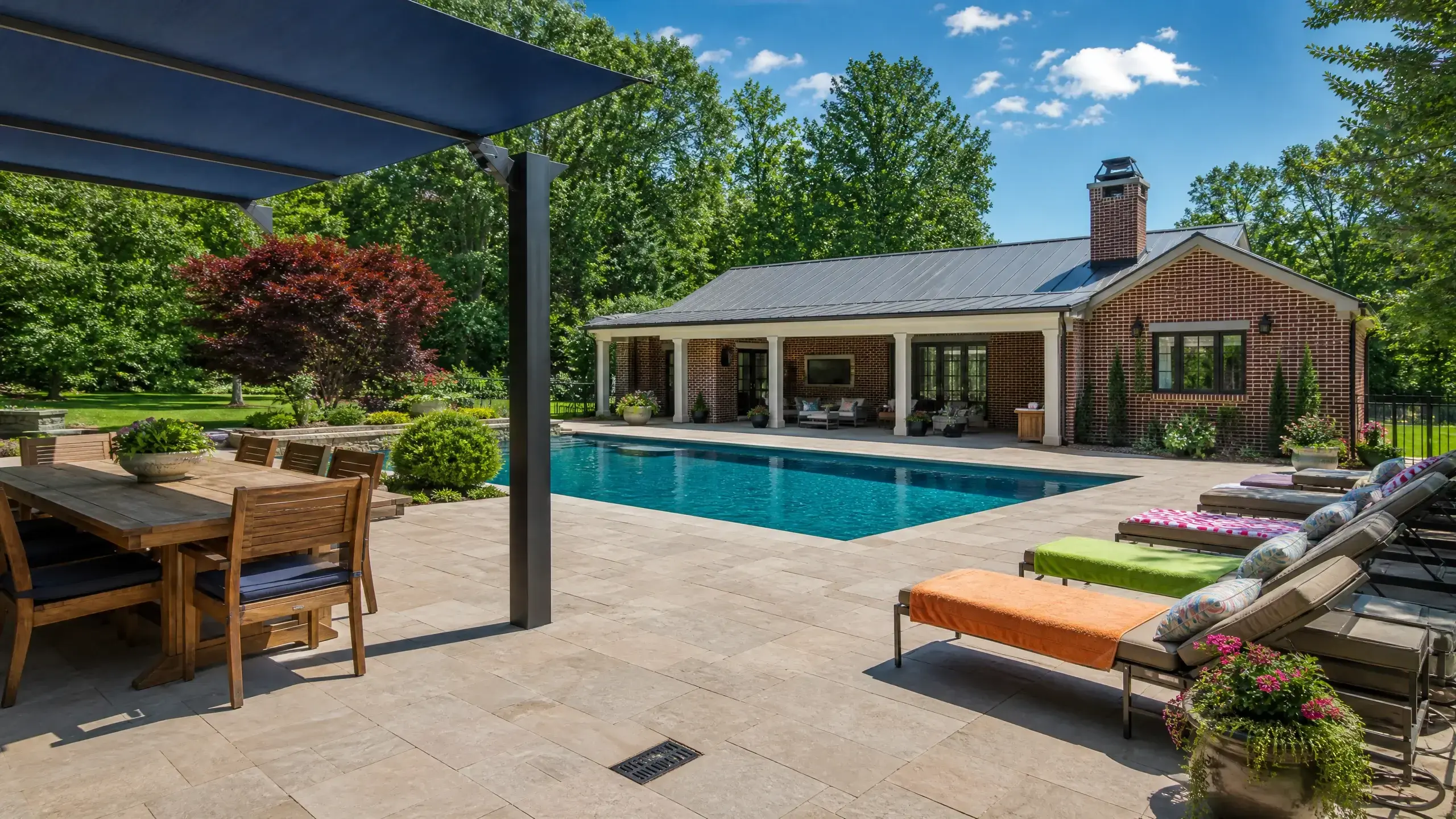 Poolside patio with lounge chairs, colorful towels, shaded dining, and a brick pool house surrounded by trees.