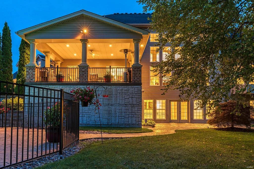 The back section of a home at dusk shows a large, covered second-story deck on a multi-level home. The house's siding is light in color, and some sections are faced with natural stone and red brick. A low wrought-iron fence surrounds part of a flat, ground-level patio. The lighting from inside the home is warm and golden, brightening the windows and the upper patio itself.