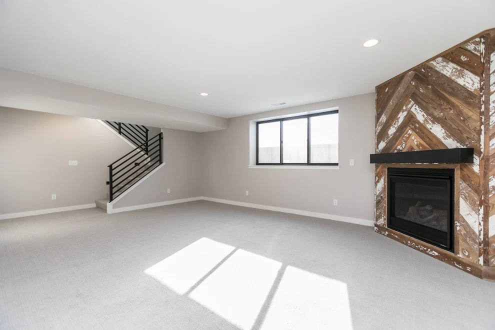 The lower level of a home with light gray walls, a white ceiling, and off-white flooring. There's a large window on the back wall, and a built-in fireplace with reclaimed wood surrounding it in the right corner of the photo. A staircase leading up from the space has a black railing.